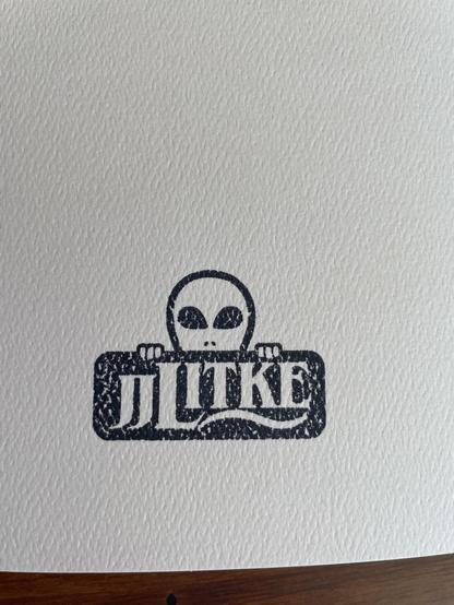 A stamped logo that reads “JJ Litke” with a little alien face peeking over the top
