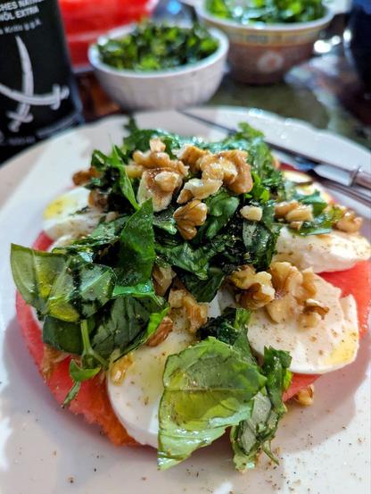 Dunkler Esstisch, weißer großer Teller mit Wassermelonenscheibe, auf der sich Mozzarellascheiben, gehackte Minze und Basilikum, Walnussstücke, Pfeffer, Meersalz und Olvenöl türmen. Besteck liegt auf dem Tellerrand. Im Hintergrund halb gefüllte Schüsseln und Teller mit den einzelnen Zutaten.