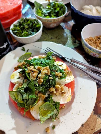 Dunkler Esstisch, weißer großer Teller mit Wassermelonenscheibe, auf der sich Mozzarellascheiben, gehackte Minze und Basilikum, Walnussstücke, Pfeffer, Meersalz und Olvenöl türmen. Besteck liegt auf dem Tellerrand. Im Hintergrund halb gefüllte Schüsseln und Teller mit den einzelnen Zutaten.