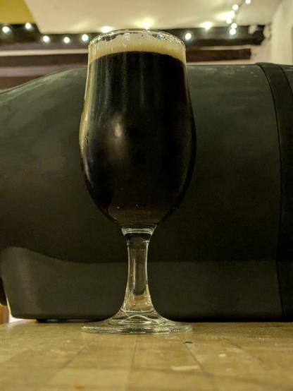 A schooner glass of very dark beer, in front of a dark grey Pinter keg.