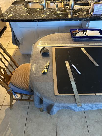 Progress photo of cutting large black silicone to counter size. It’s on flat cardboard to protect the table when I cut. White paint marker used with various metal rulers. Utility knife center next to mat has a new, sharp blade. Counter and sink show above the table.