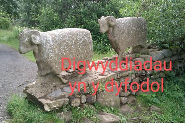 Sculpture of two grey stone sheep, one following the other, making their way along a paved path. There is green grass in the foreground and green leafy trees behind the sheep.