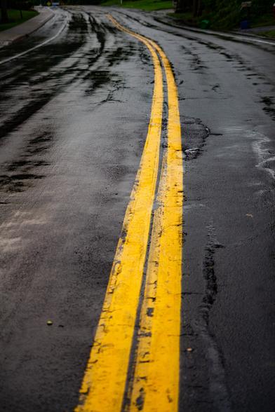 Photo of a road after rain. There are two solid yellow lines in the center curving to the left. The pavement is black and has shiny parts from the rain.
