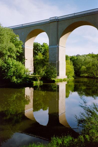Photo of a train bridge over a river, framed by greenery. The sun illuminates the arches of the bridge and their reflections in the water.