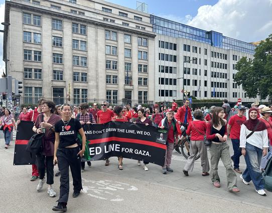 A group of red-clad people carry a banner with the main message of the red-clad demo: “If politicians won’t draw the line, we will”. It’s a black banner with a red horizontal line. Part of the text is blocked by a black clad woman with a I heart Brussels T-shirt 👕
