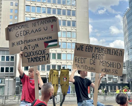 Two Flemish teenage boys are sitting on a fence showing their Dutch-language signs. One says “Damn it Netanyahu, behave yourself! I’ve got Latin exam tomorrow. Quousque Tandem?” The other says “Whoever saves a life saves all humanity. Talmud 537a, Quran 532”