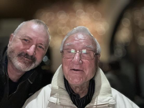 A mid-sixties man with gray hair and a beard, smiling into the camera, beside a white-haired man in his early 90's, wearing a white winter jacket. The background is bronzish bokeh.