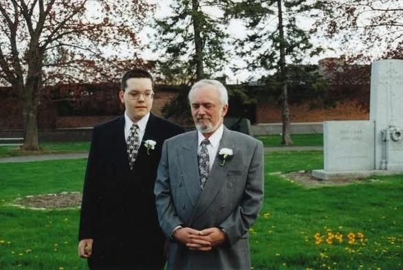 A younger me and my father, both dressed in suits with boutonnières, stand side by side in a grassy park area with trees and a monument in the background. The date on the photo reads “5-24-97,” - my parents’ wedding day (May 24, 1997).