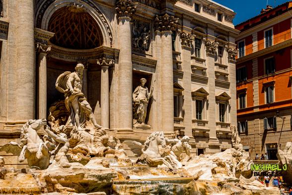A bright daytime view of Rome's Trevi Fountain, featuring elaborate marble sculptures of Oceanus, tritons, and winged horses splashing in clear water, framed by towering Baroque columns and an orange apartment building in the background.
