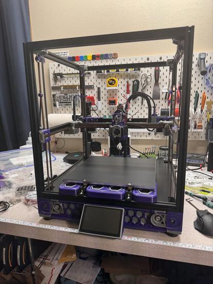 A voron 2.4 3d printer printed in purple and gray with a black frame sitting on a messy workbench with a pegboard of tools in the background. 

The printer looks nearly complete except the paneling, with a rats nest of disconnected cables around the toolhead being the primary sign that there is more work yet to be done.