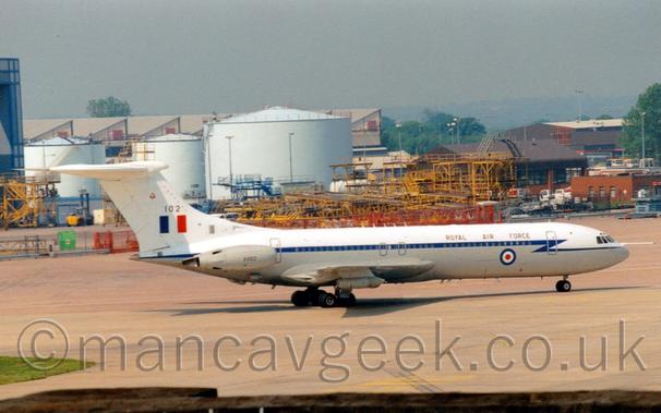 Side view of a 4 engined jet airliner taxiing from left to right.
The plane's upper surfaces are white, while the lower sections are grey, with a dark blue stripe running along the body, covering the passenger cabin windows, separating the 2 colours.
There are black "Royal Air Force" titles on the upper forward fuselage, above a large blue, white, and red roundel on the lower body.
The white tail has the large black number "102" above a blue, white, and red fin flash.
The plane's engines are mounted in pairs on the sides of the rear fuselage, slightly in front of the tail, and have the black serial "XV102" on them.
Grey concrete apron fills the foreground, with random yellow metal frames in the background in front of pale blue fuel tanks and other buildings, under blue-grey sky.