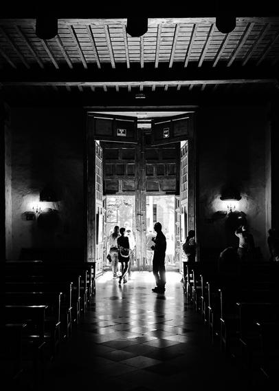 A monochrome image shows an open doorway. People are grouped at the entrance, silhouetted against bright light outside. The interior is dark and features rows of benches along the floor. The scene captures a blend of light and shadow, creating a striking contrast.