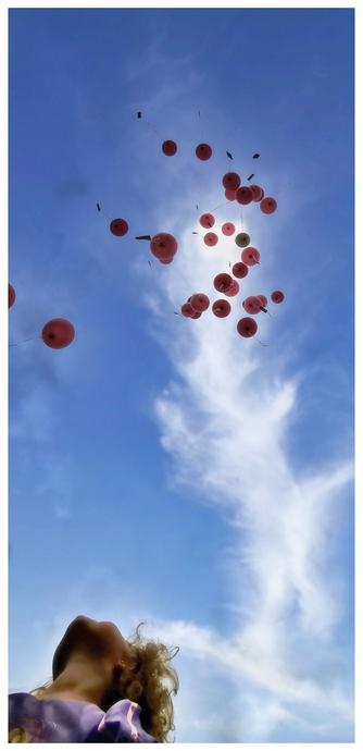 Das Bild zeigt in der linken unteren Bildecke den Oberkörper einer jungen Frau mit lockigem, blonden Haar, die mit dem Kopf nach oben in den Himmel blickt. Im Himmel schweben zahlreiche rosafarbene Ballons an dünnen Schnüren, die sich in verschiedene Richtungen ziehen. An den Enden der Schnüre sind Zettel befestigt. Der Hintergrund ist ein strahlend blauer Himmel mit zarten weißen Wolken.