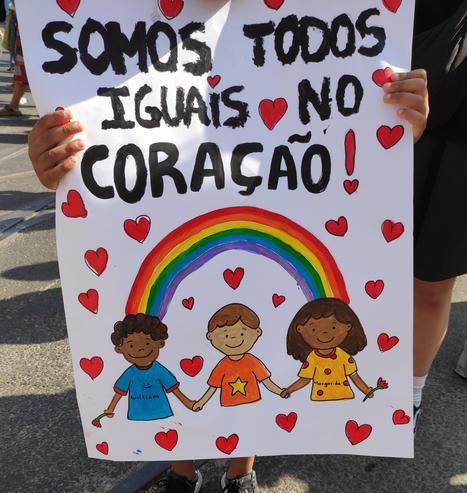 Cartaz com um desenho de três crianças de várias cores e de mãos dadas. Sobre elas, um arco-íris. AM volta delas, muitos corações. No cimo do cartaz, a frase "Somos todos iguais no Coração!"