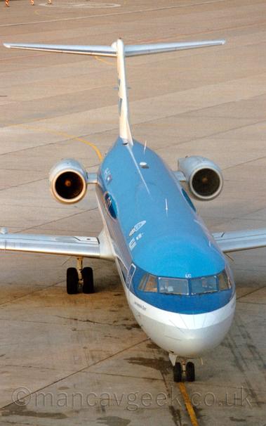 Almost head-on view from above of a twin engined jet airliner taxiing towards a spot just below the camera, following a yellow line on the apron..
The plane has blue upper surfaces, with grey lower surfaces, separated by a thick white stripe running along the body.
There are white "KLM" titles under a stylised white crown on the upper forward fuselage, a design repeated in blue on the white tail.
Grey concrete apron fills the rest of the frame.