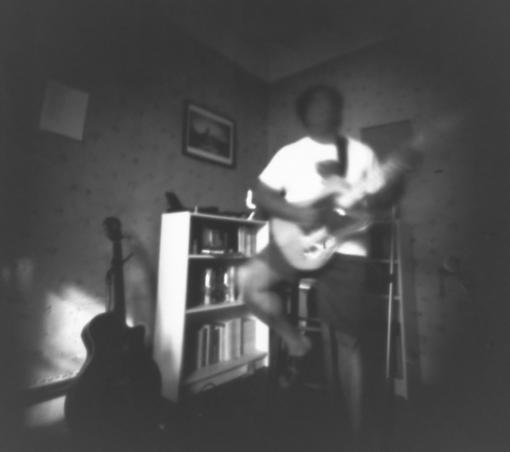 fotografía en blanco y negro. una persona apoyada en un taburete toca una guitarra eléctrica color claro. El rostro y las manos son completamente borrosos. Contra la pared hay otra guitarra y estantes con libros.