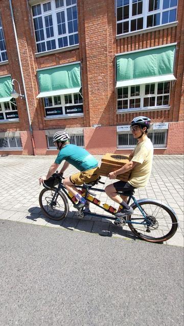 Zwei Menschen auf einem Tandem. Der hintere hält sechs Pizzakartons.