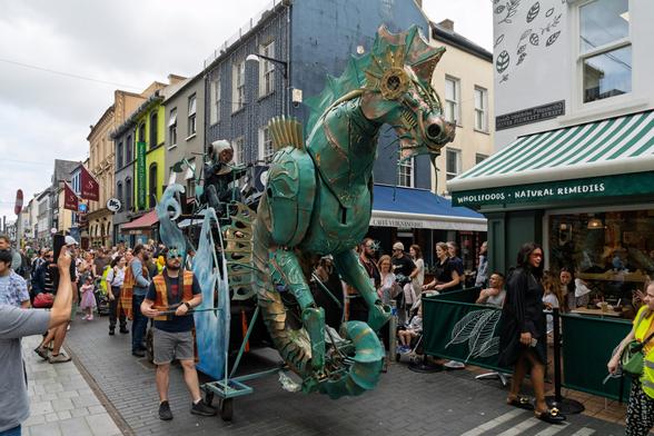 A vibrant street scene on Oliver Plunkett Street, Cork, during The Family Shenanigans parade, featuring a large, elaborate mechanical seahorse float with metallic green and gold detailing, a performer in a vintage diving suit riding the float, and crowds of onlookers lining the street. Shop signs visible include "WHOLEFOODS • NATURAL REMEDIES" and "CAFÉ VERGANO 1882".