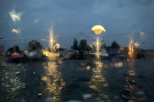 Hafenansicht am Abend durch eine nasse Scheibe fotografiert. In der Mitte eine Reihe von Booten, dahinter Lampen, deren Licht sich im Wasser in der unteren Bildhälfte spiegelt. Durch die Tropfen auf der Scheibe entstehen abstrakte Formen.
