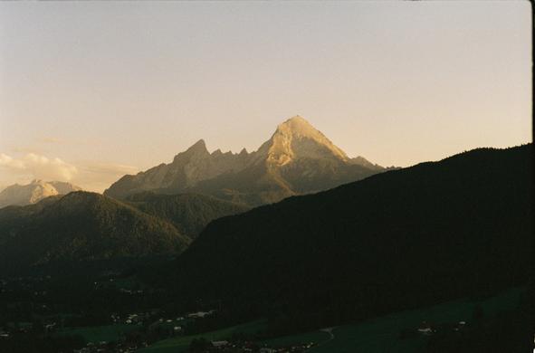 The image depicts a mountainous landscape during what appears to be either sunrise or sunset, given the warm, golden light illuminating the peak of the central mountain. The mountain in the center is prominent, with a sharp, pointed summit that is bathed in sunlight, creating a stark contrast with the darker, shadowed slopes. The surrounding mountains are less illuminated, with their slopes covered in dense green vegetation, suggesting a forested area. In the foreground, a valley is visible, with a small settlement of houses nestled among the greenery. The sky is clear with a soft gradient from light blue to a warm hue near the horizon, indicating the time of day. The overall composition of the image emphasizes the grandeur and majesty of the mountain range, with the central peak serving as the focal point.