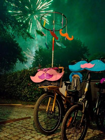 Vélos décorés avec des moustaches en carton. En fond le feu d'artifice de Tresses.