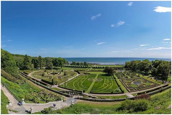 A scenic view of a landscaped garden featuring geometric patterns, trimmed hedges, and ornamental features, with the ocean visible in the background under a clear blue sky. Visitors can be seen walking along the paths.