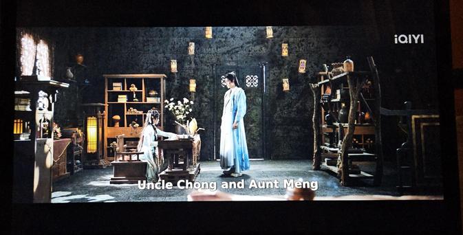 Bai Shuo writing a letter while Fan Yue looks at her.They're in a room where the walls look like painted stone and the floor look like the floor in a school hall.  While the furniture is either built out of unpainted wood or wild wood it still looks randomly placed in roughly the right places.