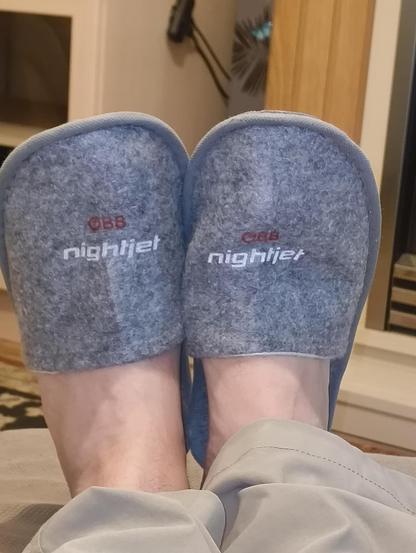 A close-up view from above shows a person wearing grey felt slippers with the "ÖBB nightjet" logo. The feet are visible, and the person is sitting