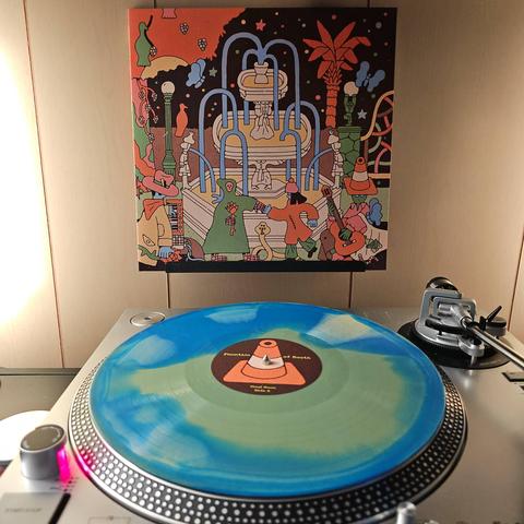 A Pangea (Mustard Yellow and Blue Merge) vinyl record sits on a turntable. Behind the turntable, a vinyl album outer sleeve is displayed. The front cover shows a multi-tiered water fountain with a grim reaper dancing with someone in front of it. A man in a cowboy hate is playing keyboard, while a creature wearing a traffice cone is playing guitar..