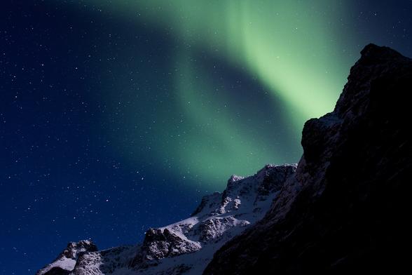 photo of a green wavy aurora against a blue sky filled with stars and framed by shadowy mountain peaks