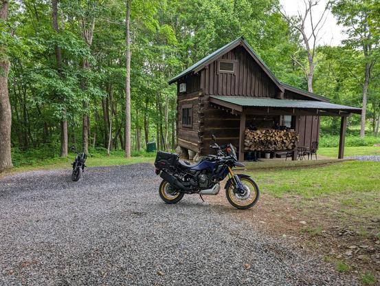A small brown cabin tucked away in the woods. Two motorcycles are parked in front of it.