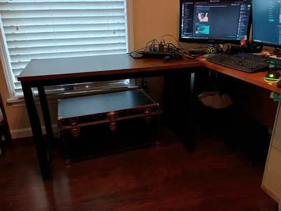 Mostly empty (and new!) desk up against the window to my home office, with another (much more cluttered) desk adjacent to it, in an L shape. There is an older steamer trunk underneath the new desk.