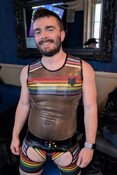 A person Wearing a transparent Latex top and chap shorts combo.
both items are decorated with ines of latex in the pride flag colours