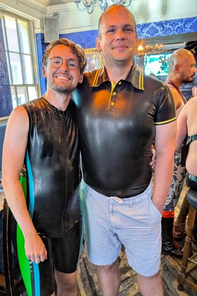 Two people, one in a black latex surfsuit with green and blue piping. the other in a black latex polo shirt with yellow piping.