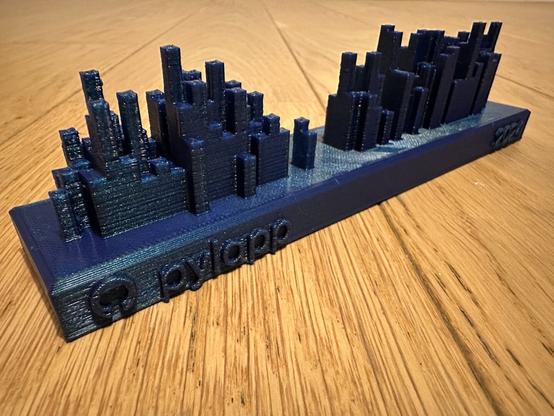 A 3D-printed GitHub commits history for user “pylapp” on year 2024. These string used is blue with some foil effect. The object is on a wood surface ans is in fact a 3D bar charts showing commits history on GitHub platform. The logo of GitHub is also displayed left to the “pylapp” username. The bare look like buildings or skyscrapers. The higher it is the higher was the number of commits for the given day. In the middle of the object there is a hole, maybe for months May and June.