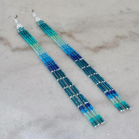 A pair of long beaded dangle earrings in shades of blue, turquoise, and white, on a marble background.