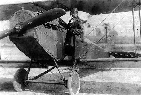 A young black woman standing on the wheel of an early propeller plane. She's wearing a flying uniform of trousers, boots, a long leather coat and a leather helmet.