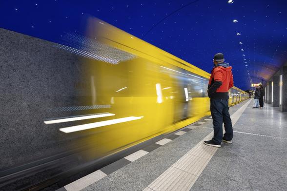 Slow motion shot of a yellow metro train entering the platform at Museumsinsel U-Bahn Station in Berlin, under a painted dark blue starry ceiling. A passenger in a bright red jacket is waiting in the foreground on the platfom adding some more color contrast to the bright yellow and dark blue tones.