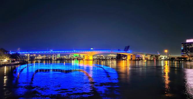 Late night view over a vast, calm, dark river with bright blue bridge lighting reflecting upon the water's surface below.