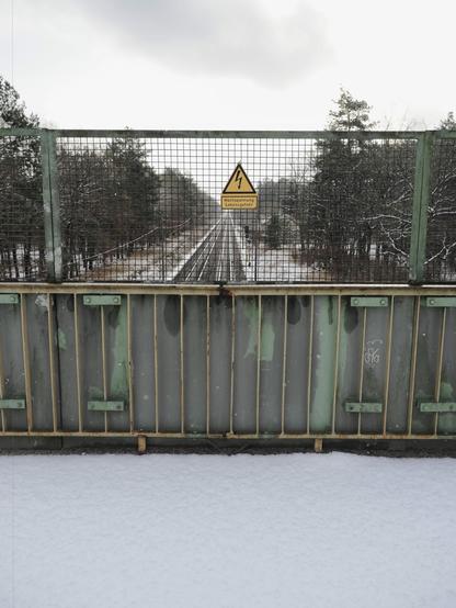 A metal fence with a warning sign displaying a lightning bolt symbol and German text "Hochspannung Lebensgefahr" (High voltage danger to life). Below the fence is a weathered green metal railing. Beyond the fence, a railway track extends into the distance, flanked by snow-covered ground and trees. Overcast sky above.