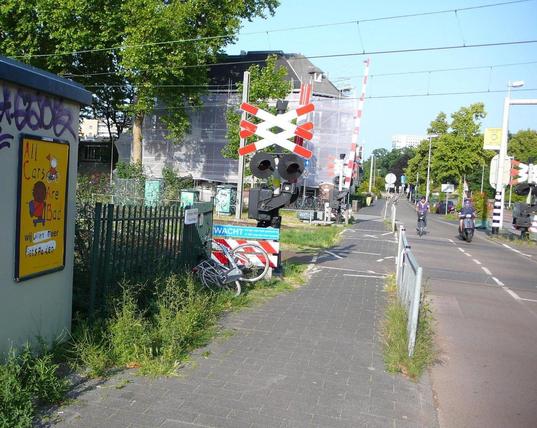 spoorwegovergang met rood/witte waarschuwingsborden, een fietser en brommer die overkomen, een pand, wat bomen en een muurtje lins met het bewerkte reclamebord