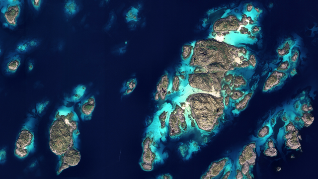A satellite picture of scattered rocky islands with patches of greenery, surrounded by deep navy water and vivid turquoise shallows and reefs.