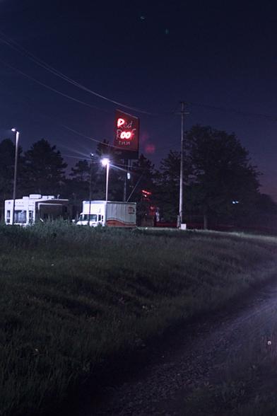 An illuminated Red Roof Inn sign beside a highway below a dark night sky, with a truck and street lights nearby. The sign originally speled out the name of the motel in red neon letters. All letters are still visible but most are dead, plus part of the "R." It now says "Poo."