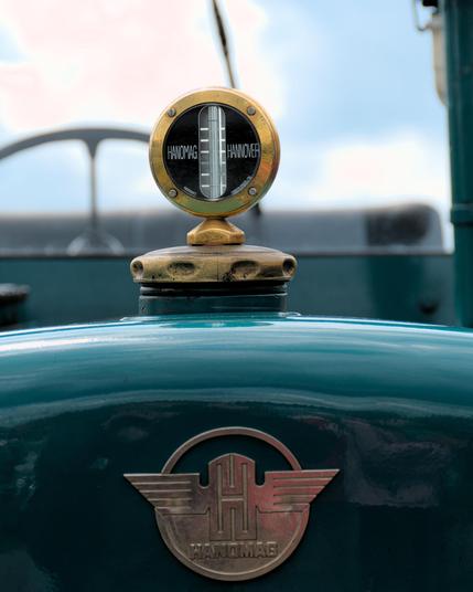 Circular brass radiator cap with thermometer, labeled "HANOMAG" on a teal vehicle. Below, the Hanomag emblem featuring a stylized "H" with wings. Blurry background includes sky and vehicle elements.