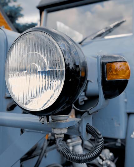 Close-up of the front headlight and blinker of a vintage blue vehicle. The large, round headlight features a textured glass surface, while the smaller amber blinker is mounted above it on the right. Power cables and mounting brackets are visible, with parts of the vehicle’s frame and windshield wipers in the background.