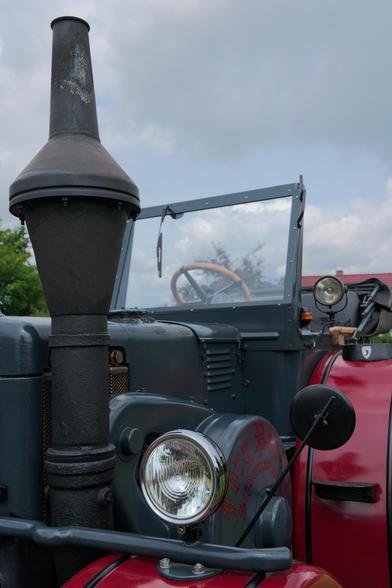 Antique vehicle with a large black chimney-like structure on the left. The front features a round headlight and a badge with stylized text. A side mirror is attached near the front. The body is dark with red accents. The driver's seat with a wooden steering wheel is visible behind a windshield. Cloudy sky and some greenery in the background.
