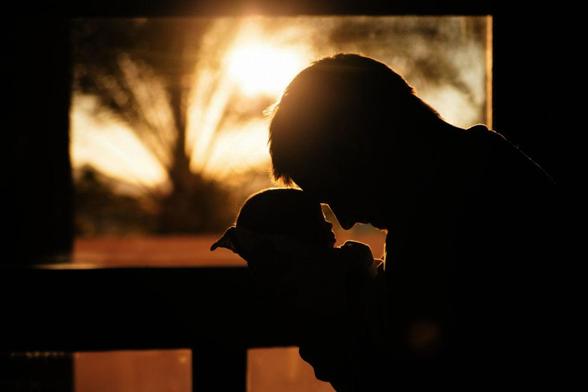 A silhouette of a man holding a baby, touching his forehead to the infant's. The room they are in is completely shadowed and the sun is rising golden through a window behind them.