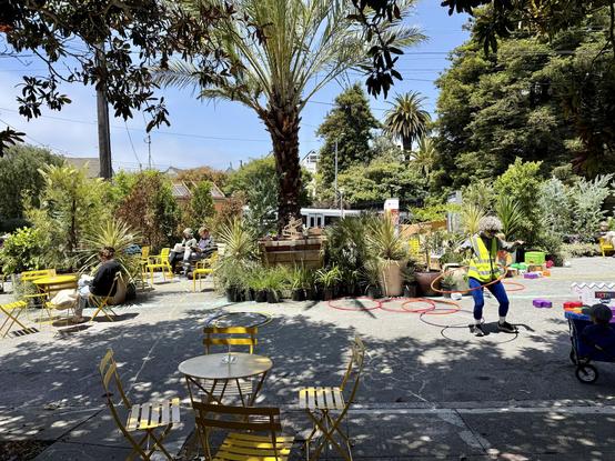 Photograph of an urban street closed to traffic and outfitted with hundreds of potted plants and trees, yellow cafe tables and chairs, and physical playthings including #HulaHoop rings and giant #Tetris blocks. Several people are seated or hula hooping in clear and dappled sunlight, while a light rail train is partially visible in the background behind the parklet.