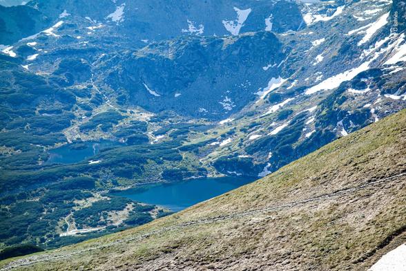 Looking down a mountainside at two snow-fed lakes perched in a valley below and between peaks. There a patches of snow still remaining although it is mid-June. There mountainsides are mostly bare on the steeper slopes, but there are stands of trees around the lakes and in the valley.