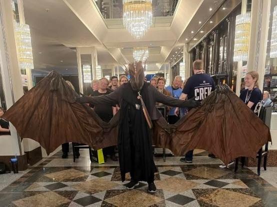 A person in a dramatic dragon costume with large, outstretched wings stands in a stylish venue, surrounded by a group of people. The setting features elegant lighting and decor.